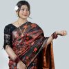 A woman looking down and posing elegantly against a plain white studio background. She is wearing a premium black silk saree featuring large, bold abstract floral patterns in crimson red and terracotta orange inspired by Jamdani designs. The saree features a high-shine copper-red silk inner lining. She has styled it with a black lace sleeve blouse, a delicate gold necklace, maroon bangles, and a fresh white plumeria flower in her hair.