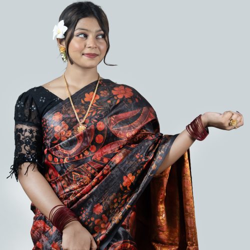 A woman looking down and posing elegantly against a plain white studio background. She is wearing a premium black silk saree featuring large, bold abstract floral patterns in crimson red and terracotta orange inspired by Jamdani designs. The saree features a high-shine copper-red silk inner lining. She has styled it with a black lace sleeve blouse, a delicate gold necklace, maroon bangles, and a fresh white plumeria flower in her hair.
