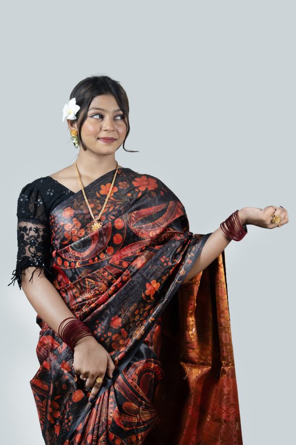 A woman looking down and posing elegantly against a plain white studio background. She is wearing a premium black silk saree featuring large, bold abstract floral patterns in crimson red and terracotta orange inspired by Jamdani designs. The saree features a high-shine copper-red silk inner lining. She has styled it with a black lace sleeve blouse, a delicate gold necklace, maroon bangles, and a fresh white plumeria flower in her hair.