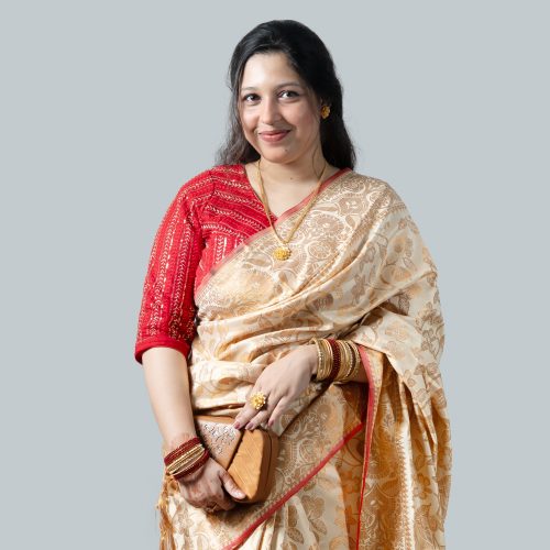 A woman smiling and posing against a plain light gray studio background while holding a single red rose. She is wearing a premium off-white and cream Katan silk saree detailed with heavy, intricate golden zari floral embroidery all over. She has styled it with a contrasting bright red sequined blouse, gold necklaces, a large gold ring, and red bangles.