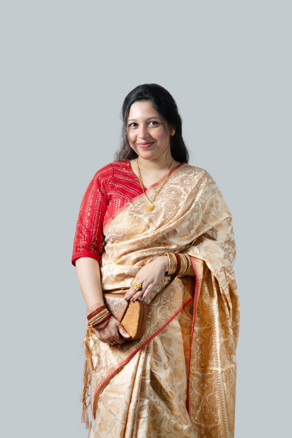 A woman smiling and posing against a plain light gray studio background while holding a single red rose. She is wearing a premium off-white and cream Katan silk saree detailed with heavy, intricate golden zari floral embroidery all over. She has styled it with a contrasting bright red sequined blouse, gold necklaces, a large gold ring, and red bangles.
