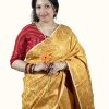 A woman sitting and smiling gently against a plain light gray studio background. She is wearing a premium mustard yellow Katan silk saree covered in intricate, woven golden paisley and floral patterns. She has styled the saree with a contrasting bright red blouse featuring gold embroidery, a gold necklace with a large pendant, matching gold earrings, and a stack of red and gold bangles.