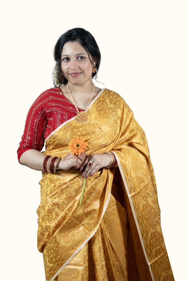 A woman sitting and smiling gently against a plain light gray studio background. She is wearing a premium mustard yellow Katan silk saree covered in intricate, woven golden paisley and floral patterns. She has styled the saree with a contrasting bright red blouse featuring gold embroidery, a gold necklace with a large pendant, matching gold earrings, and a stack of red and gold bangles.