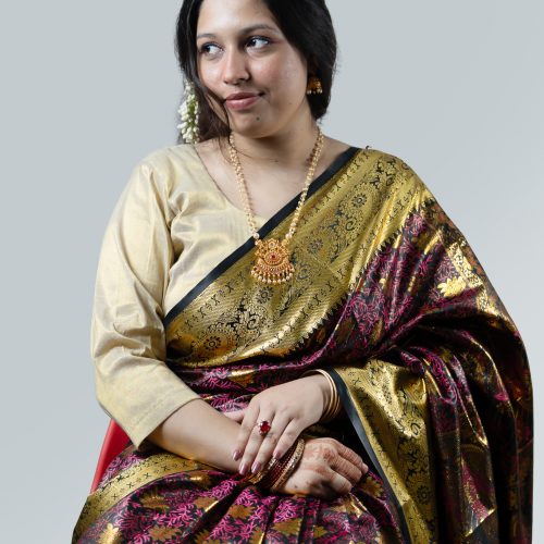 A professional studio product photograph from yourfashionbd.xyz showcasing a woman modeling a premium midnight black and scarlet red Katan silk saree. The saree features intricate, continuous red floral and paisley Minakari thread embroidery across the dark body, cascading into a high-luster amber-gold inner lining and detailed border. She is styled with a black lace-detailed short-sleeve blouse, a white flower tucked into her bun, red bangles, and a delicate gold necklace.