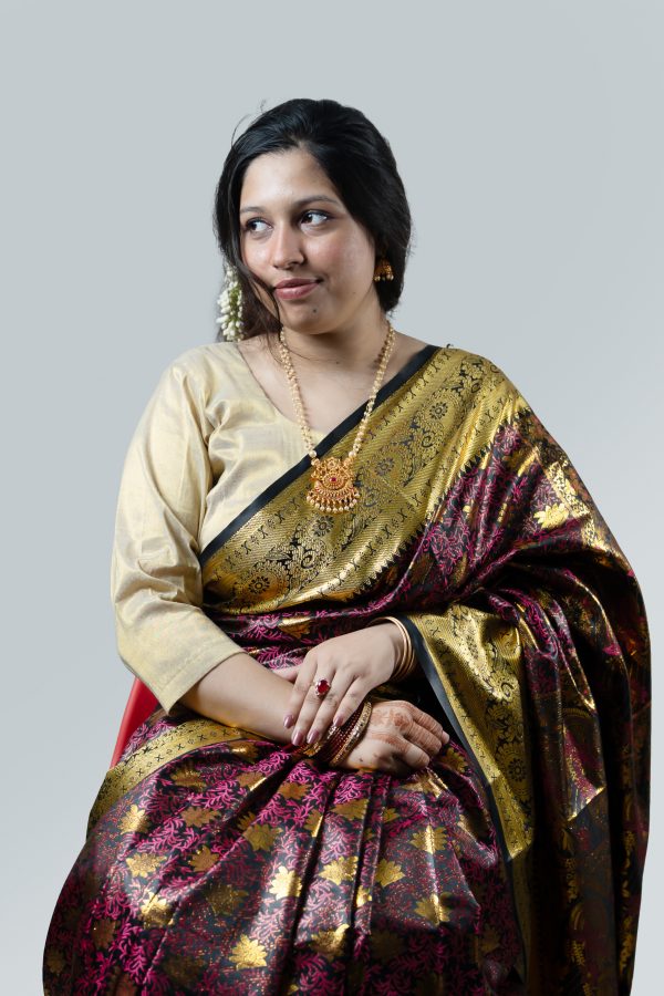 A professional studio product photograph from yourfashionbd.xyz showcasing a woman modeling a premium midnight black and scarlet red Katan silk saree. The saree features intricate, continuous red floral and paisley Minakari thread embroidery across the dark body, cascading into a high-luster amber-gold inner lining and detailed border. She is styled with a black lace-detailed short-sleeve blouse, a white flower tucked into her bun, red bangles, and a delicate gold necklace.